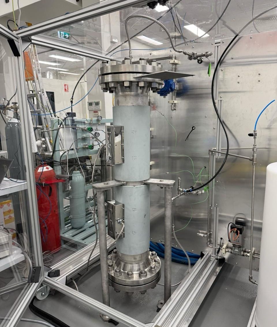 A metal tube with large discs clamped at each end and held in a metal frame. Gas cylinders in the background give an impression of scale which is about 1.5 to 2 metres tall and 20 or 30 centimetres wide.