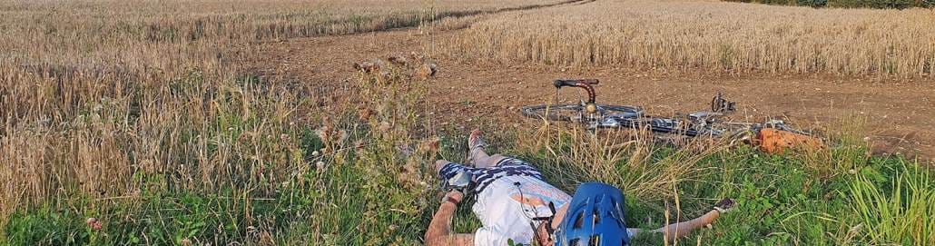The image shows a person lying prone in a field, bicycle straps dangling in the grass, with tall, dry grasses swaying in the background.
AI-generated content may be incorrect.