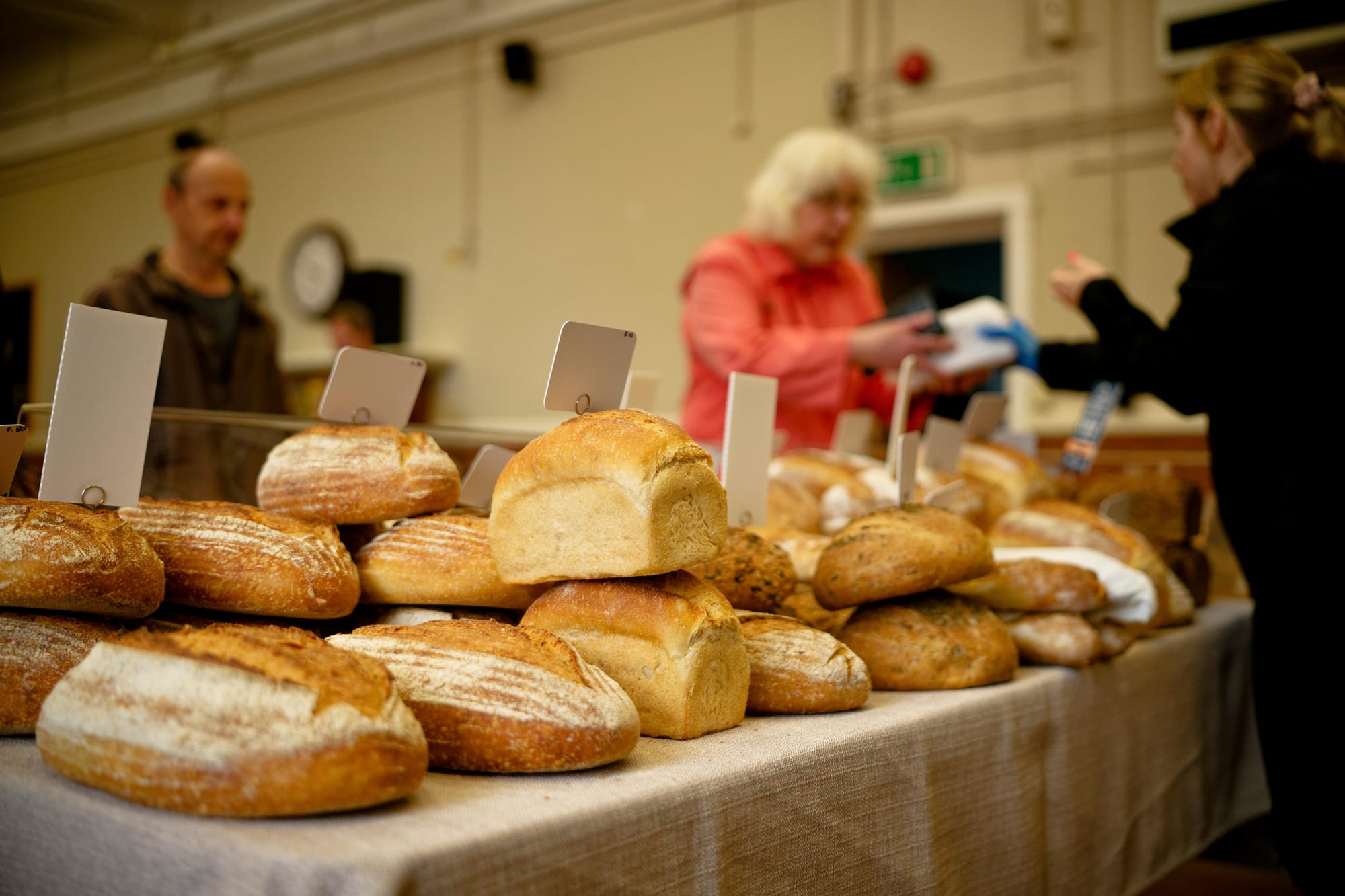 Loaves of bread at a bread stall, with stall holder handing over a wrapped loaf to a customer, with another customer waiting behind