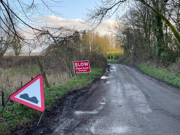 A country road, with signs 'Uneven Road ahead' warning triangle and 'SLOW Failed Road Surface'